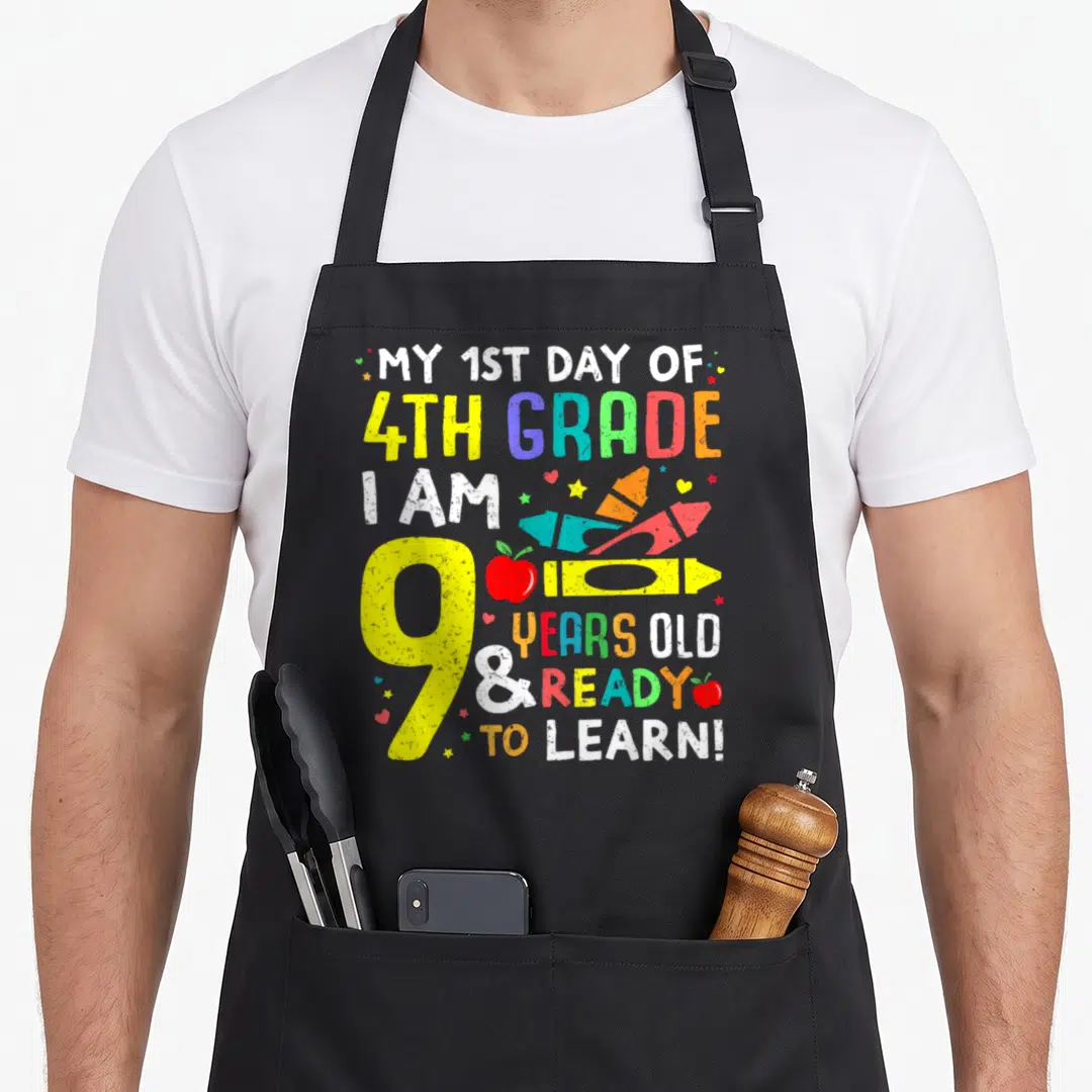4th school grade apron - Stacked text with yellow pencil, red apple, rockets, hearts, stars in yellow red blue green — shown on a black cotton canvas apron against a white background