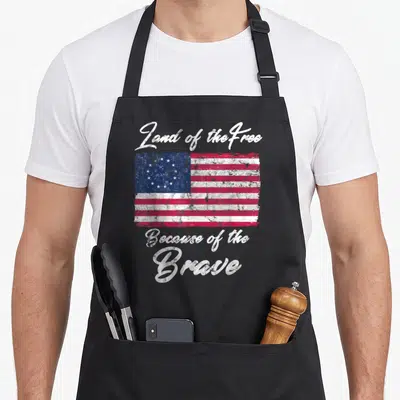 Betsy patriotic aprons - Distressed red white blue flag with Land of the Free Because of the Brave text — shown on a black cotton canvas apron against a white background