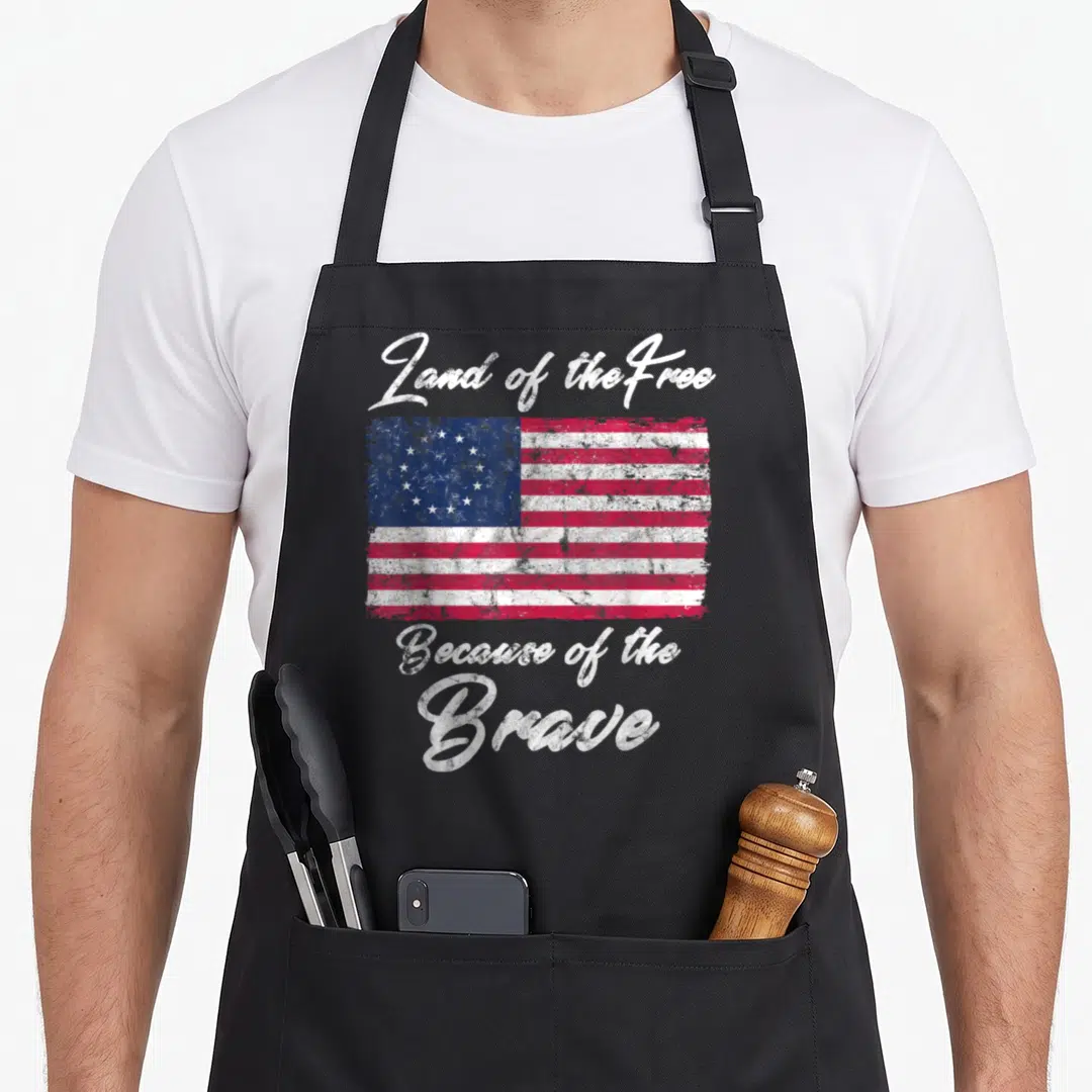 Betsy patriotic aprons - Distressed red white blue flag with Land of the Free Because of the Brave text — shown on a black cotton canvas apron against a white background