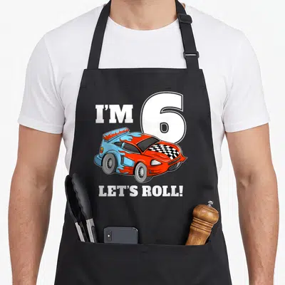 Six car apron - I'M 6, LET'S ROLL! text above and below a red blue and orange cartoon race car with a checkered hood — shown on a black cotton canvas apron against a white background