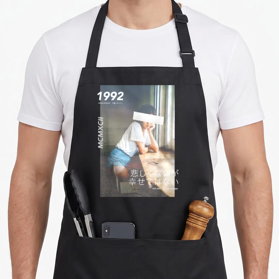 Funny japanese streetwear apron - 1992 woman photo with NATHAN KAWANISHI and not sad not happy text — shown on a black cotton canvas apron against a white background