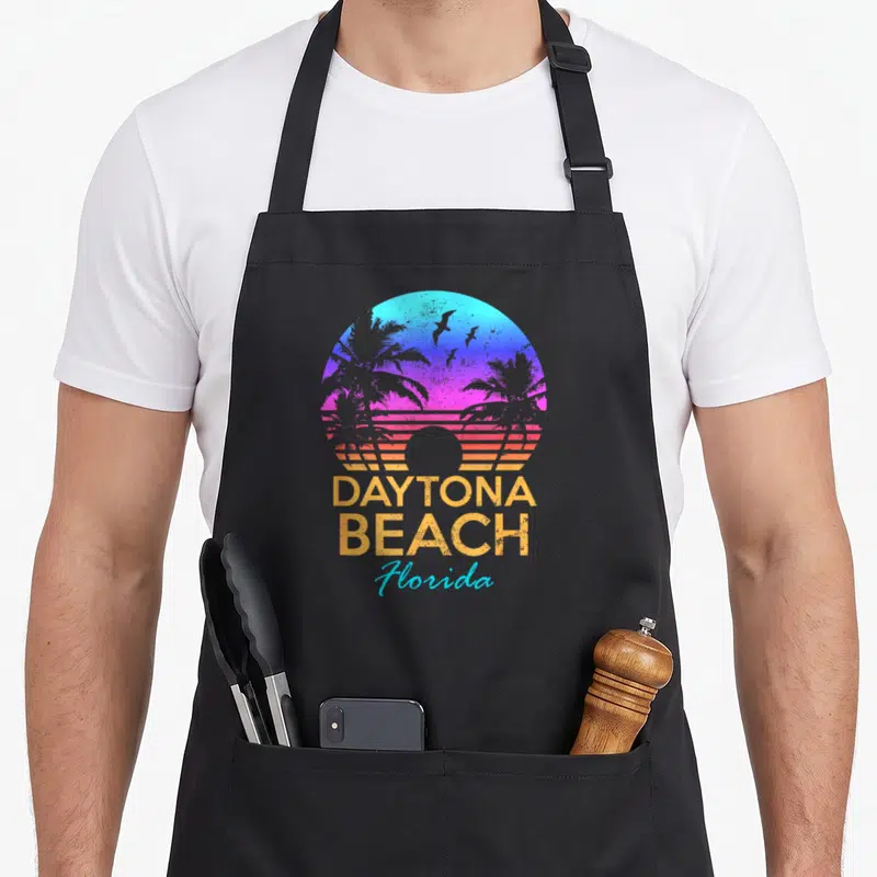Daytona summer aprons - Retro circular sun with palm trees, birds, and DAYTONA BEACH Florida text in blue purple pink — shown on a black cotton canvas apron against a white background