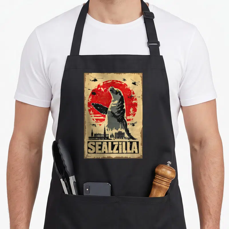 Sealzilla birthday apron - Vintage grey seal towering over city skyline with red sun and helicopters — shown on a black cotton canvas apron against a white background