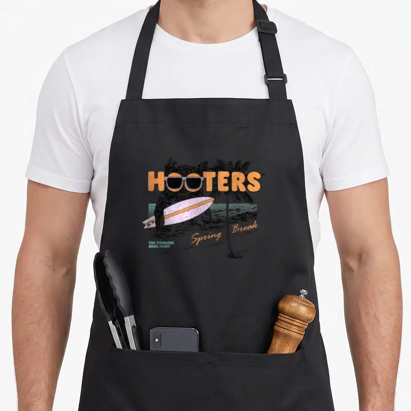 Hooter spring aprons - Retro owl wearing sunglasses holding a surfboard with Hooters text and palm tree — shown on a black cotton canvas apron against a white background