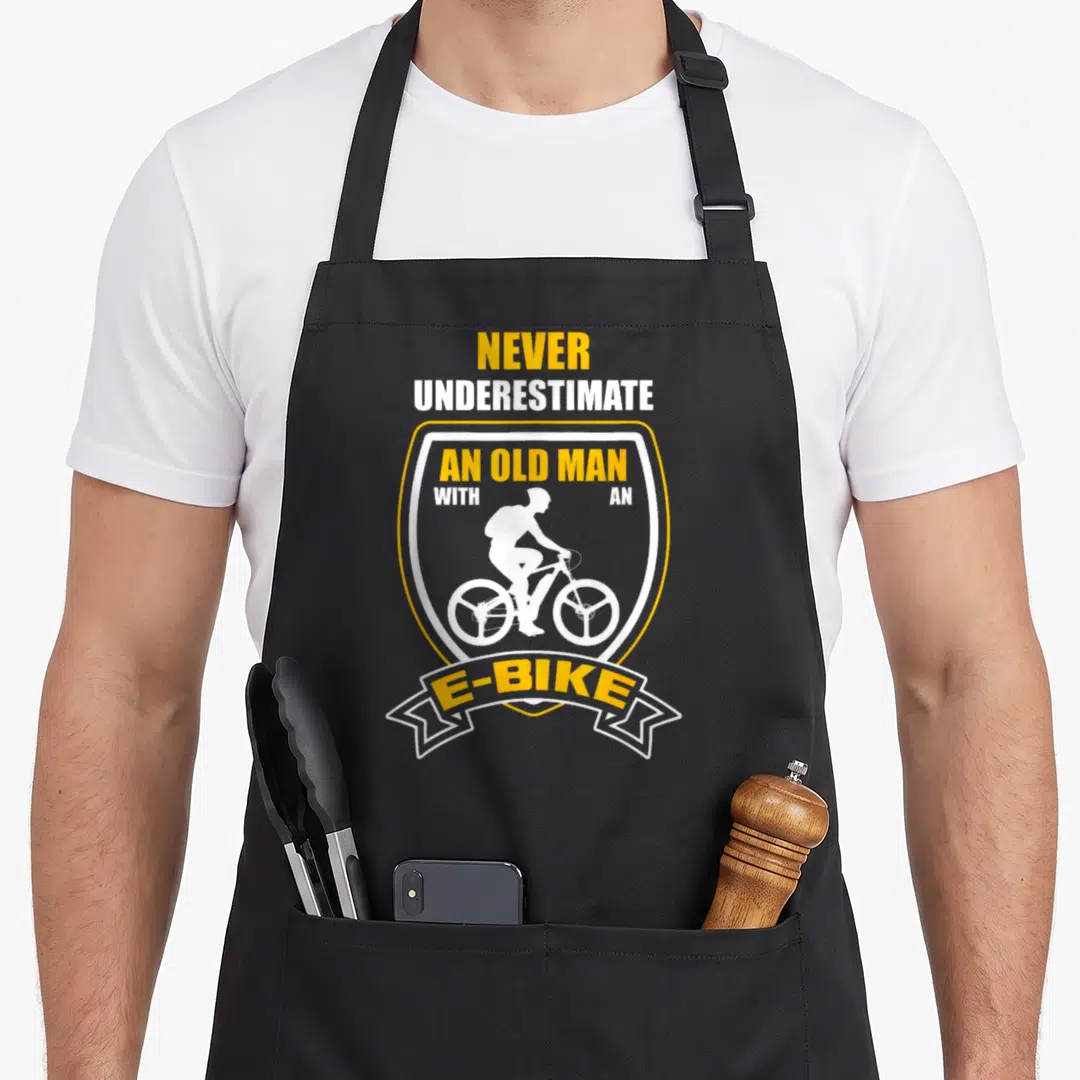 Underestimate ebike bike apron - Yellow and white shield with old man e-bike silhouette and stacked text — shown on a black cotton canvas apron against a white background
