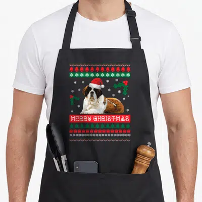 Bernard ugly christmas sweater apron - Saint Bernard wearing Santa hat surrounded by bells snowflakes trees and MERRY — shown on a black cotton canvas apron against a white background