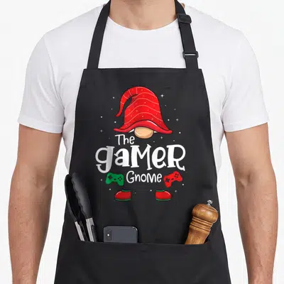 Gamer christmas gnome apron - Cartoon gnome with red hat, white text, two game controllers, and small stars scattered — shown on a black cotton canvas apron against a white background