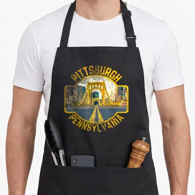 Funny pittsburgh pennsylvania girl apron - Yellow framed bridge and skyline with PITTSBURGH PENNSYLVANIA text in retro — shown on a black cotton canvas apron against a white background