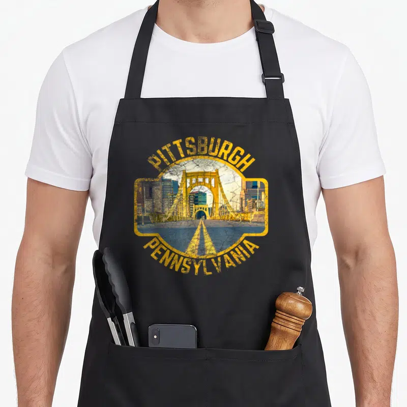 Funny pittsburgh pennsylvania girl apron - Yellow framed bridge and skyline with PITTSBURGH PENNSYLVANIA text in retro — shown on a black cotton canvas apron against a white background