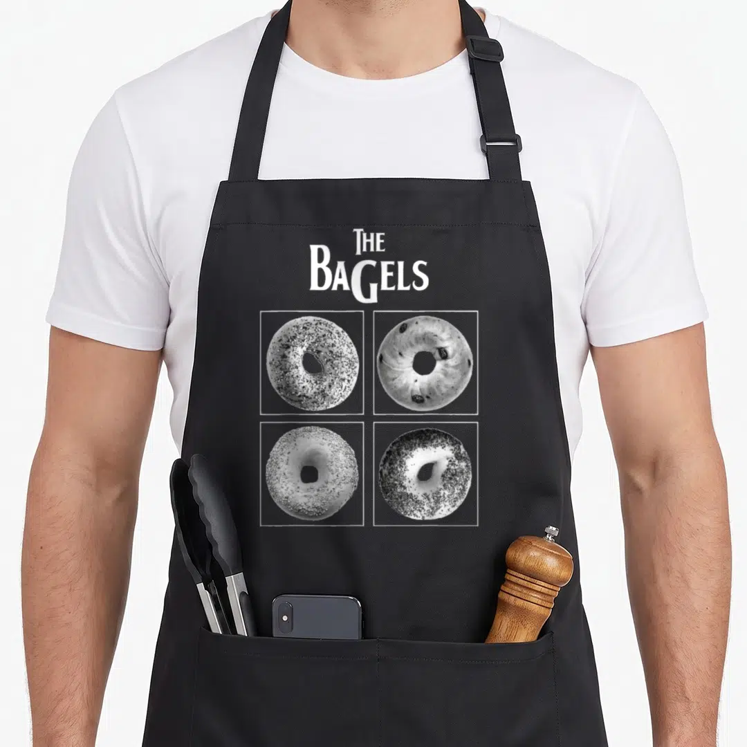 Bread food aprons - THE BAGELS text above four bagels in square frames, grayscale — shown on a black cotton canvas apron against a white background
