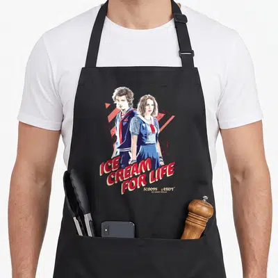 Steve ice cream apron - Two cartoon figures in blue uniforms with red text reading ICE CREAM FOR LIFE and geometric — shown on a black cotton canvas apron against a white background