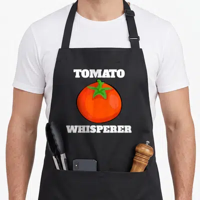 Tomato gardener apron - TOMATO WHISPERER text above and below red cartoon tomato with green stem — shown on a black cotton canvas apron against a white background