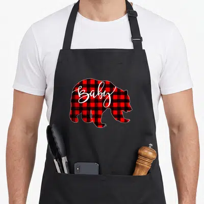 Baby buffalo plaid christmas apron - Bear silhouette filled with red black plaid and white script Baby text — shown on a black cotton canvas apron against a white background