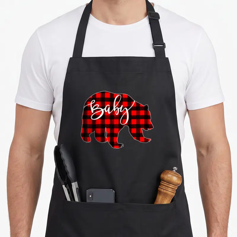 Baby buffalo plaid christmas apron - Bear silhouette filled with red black plaid and white script Baby text — shown on a black cotton canvas apron against a white background