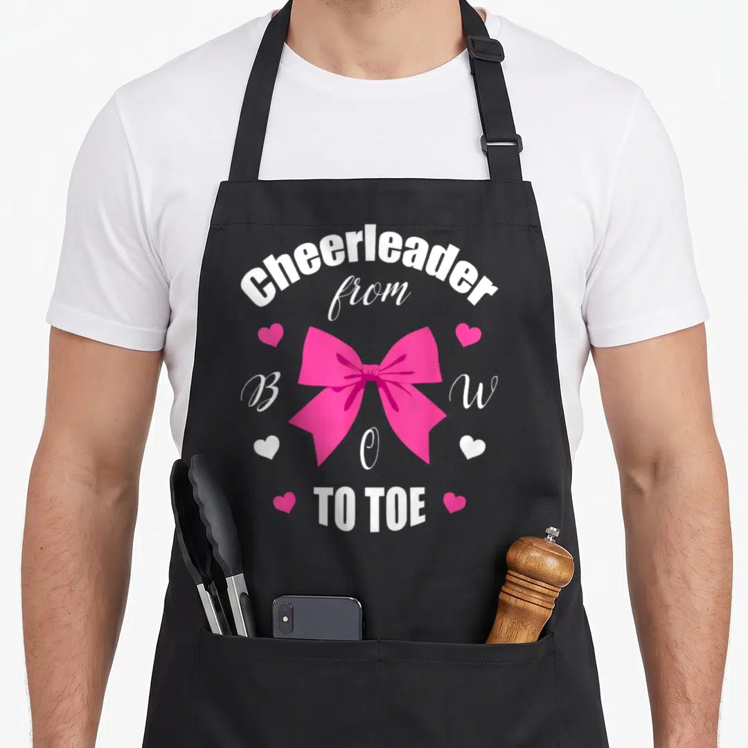 Cheerleader bow apron - Pink bow surrounded by white text Cheerleader from Bow to Toe and four hearts — shown on a black cotton canvas apron against a white background