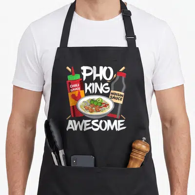 Asia pho apron - Cartoon pho bowl flanked by red chili and black hoisin sauce bottles with white PHO KING AWESOME text — shown on a black cotton canvas apron against a white background