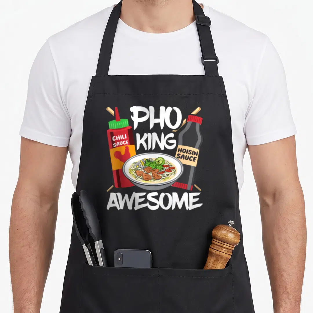 Asia pho apron - Cartoon pho bowl flanked by red chili and black hoisin sauce bottles with white PHO KING AWESOME text — shown on a black cotton canvas apron against a white background