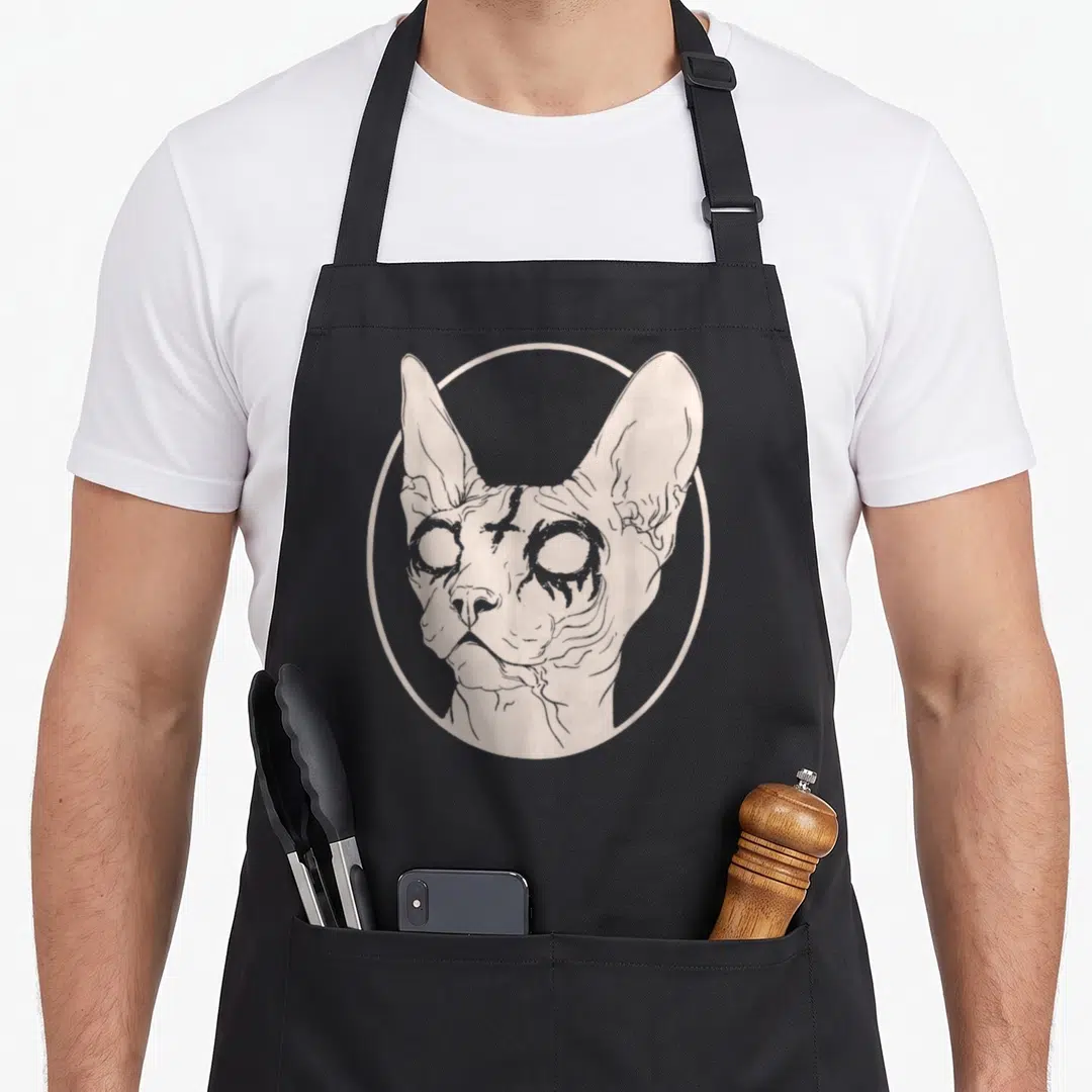 Satanic cat apron - Beige line art of a sphynx cat head with hollow eyes inside a circle — shown on a black cotton canvas apron against a white background