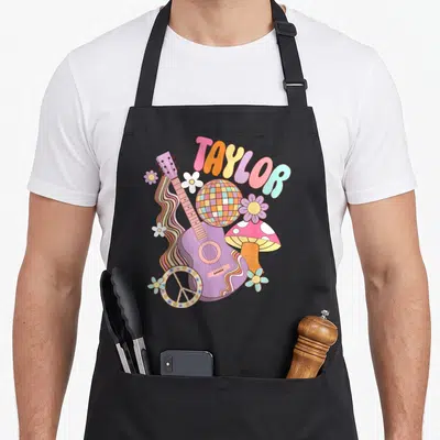Retro taylor 80s apron - TAYLOR text over purple guitar, disco ball, mushroom, flowers, and peace symbol — shown on a black cotton canvas apron against a white background
