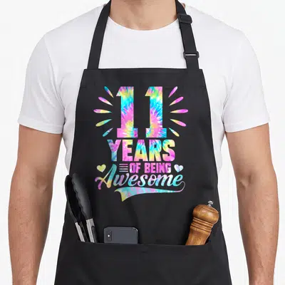 11th tie apron - Tie-dye 11 years of being awesome text with hearts and sunburst rays in pink blue yellow purple — shown on a black cotton canvas apron against a white background