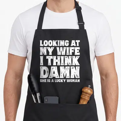 Joke husband apron - White stacked text reading LOOKING AT MY WIFE I THINK DAMN SHE IS A LUCKY WOMAN — shown on a black cotton canvas apron against a white background