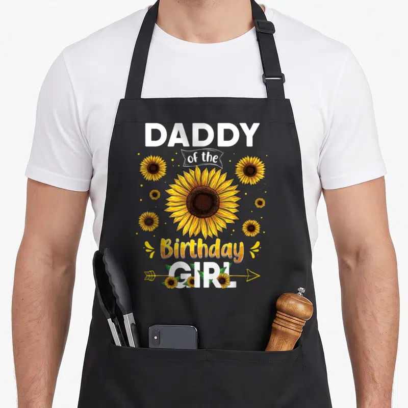 Daddy sunflower apron - Cartoon sunflower with Daddy of the Birthday Girl text and arrow — shown on a black cotton canvas apron against a white background