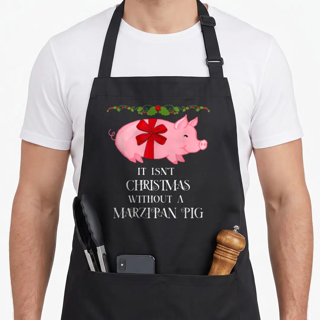 Norwegian scandinavian aprons - Pink cartoon pig with red ribbon and white text below a green holly vine border — shown on a black cotton canvas apron against a white background