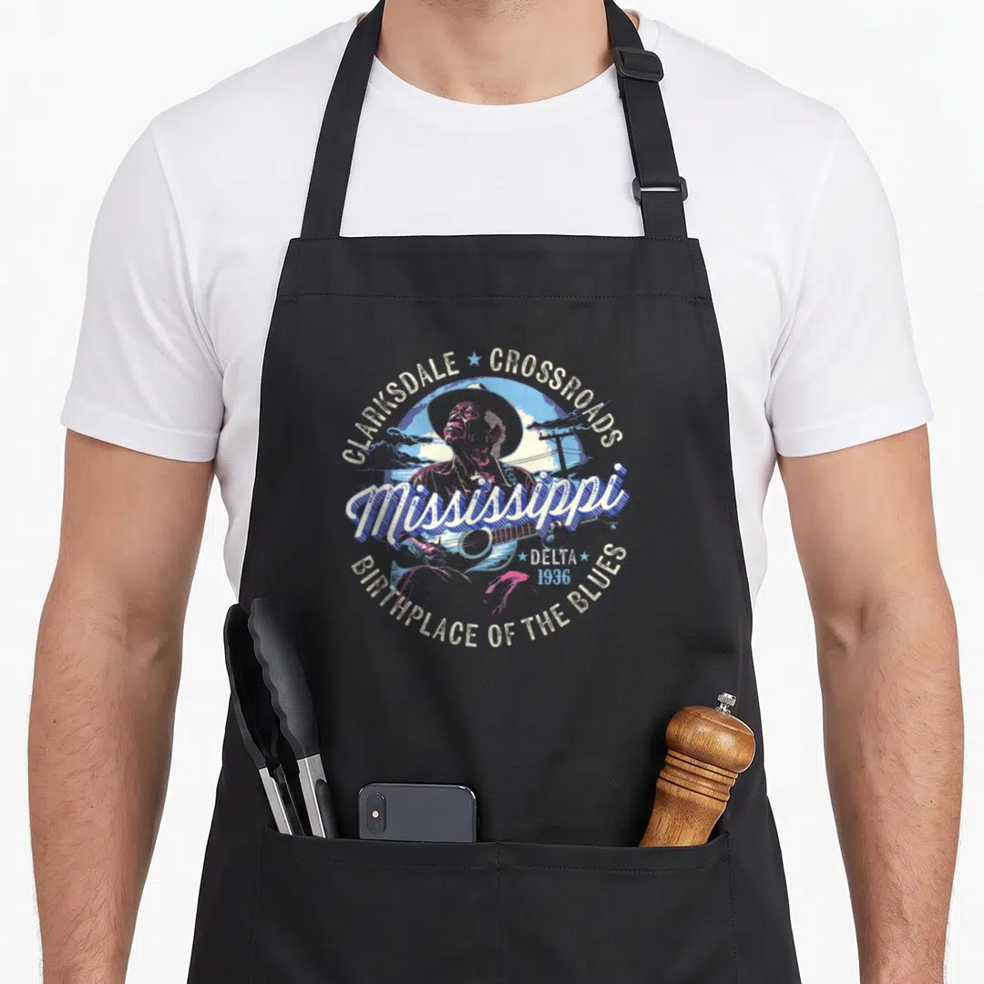 Clarksdale mississippi apron - Retro print of man playing guitar with Clarksdale Crossroads text in blue and purple — shown on a black cotton canvas apron against a white background