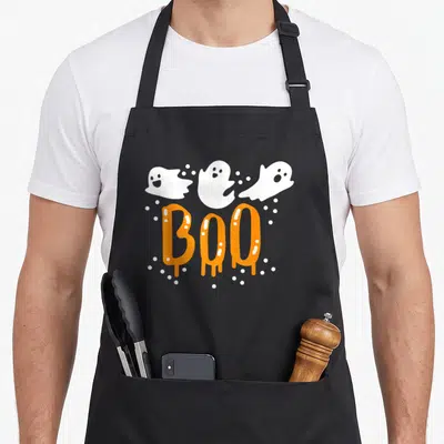 Retro ghost halloween apron - Three white cartoon ghosts above dripping orange BOO text surrounded by white dots — shown on a black cotton canvas apron against a white background
