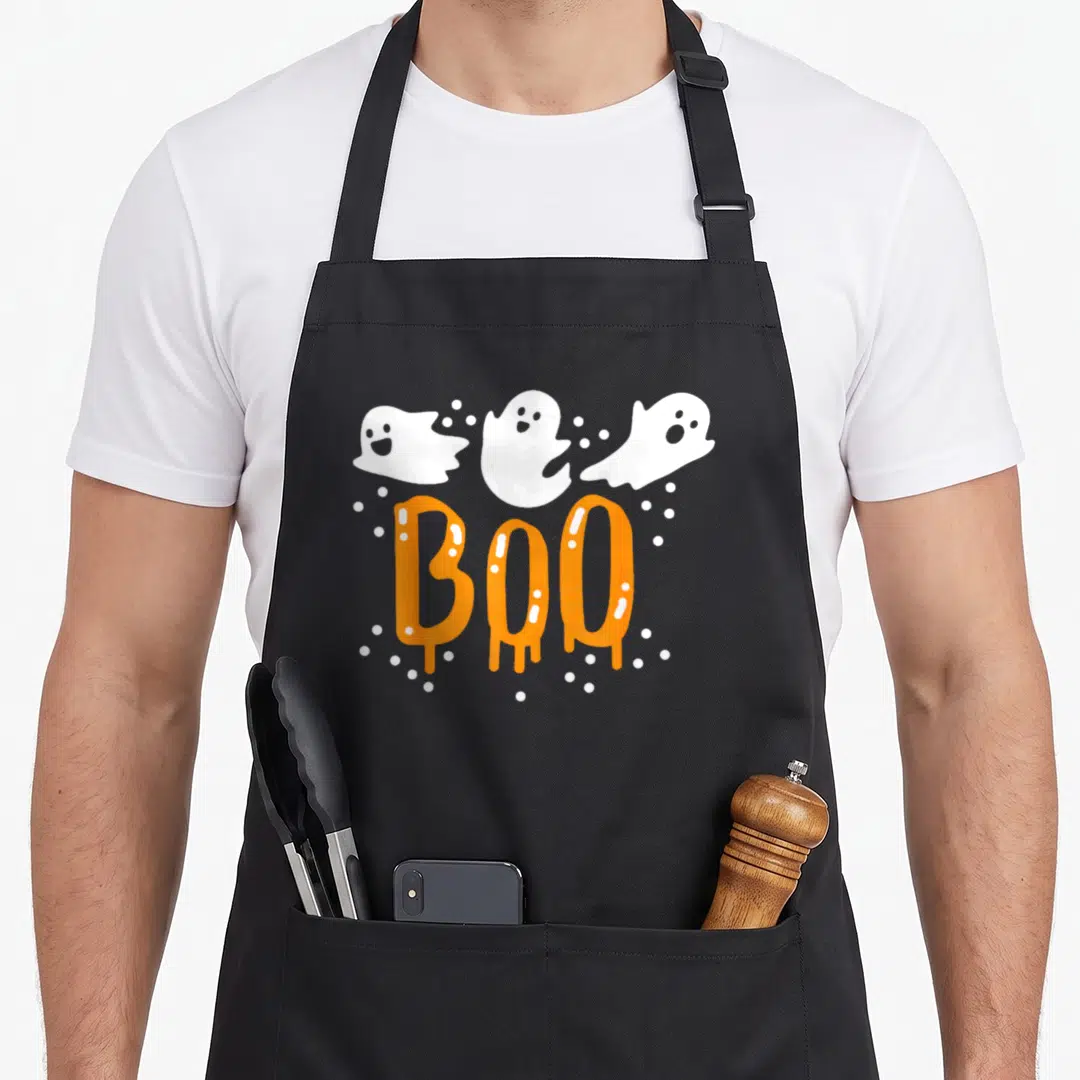 Retro ghost halloween apron - Three white cartoon ghosts above dripping orange BOO text surrounded by white dots — shown on a black cotton canvas apron against a white background