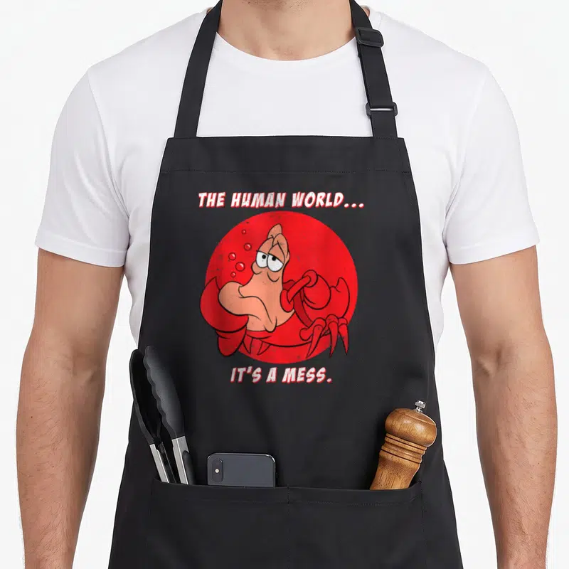 Sebastian mermaid apron - Crab inside red circle with text THE HUMAN WORLD IT'S A MESS — shown on a black cotton canvas apron against a white background