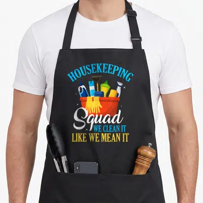 Clean housekeeper apron - Cartoon bucket with supplies and text HOUSEKEEPING SQUAD WE CLEAN IT LIKE WE MEAN IT in blue — shown on a black cotton canvas apron against a white background
