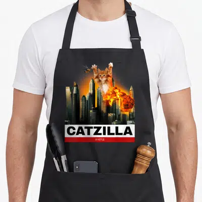 Catzilla halloween apron - Giant orange cat towers over city skyscrapers with explosion and helicopter above CATZILLA — shown on a black cotton canvas apron against a white background