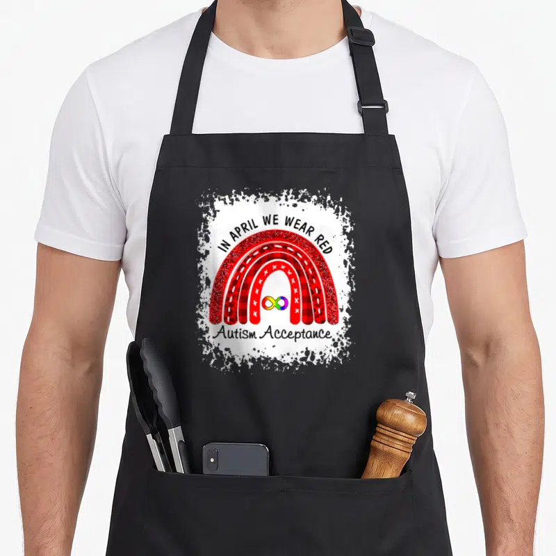 Autism acceptance april apron - Red patterned rainbow with infinity symbol and 'In April We Wear Red' text — shown on a black cotton canvas apron against a white background