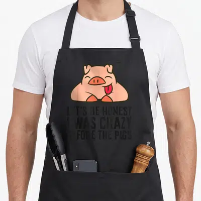 Honest crazy pig man apron - Pink cartoon pig above black text LET'S BE HONEST I WAS CRAZY BEFORE THE PIGS — shown on a black cotton canvas apron against a white background