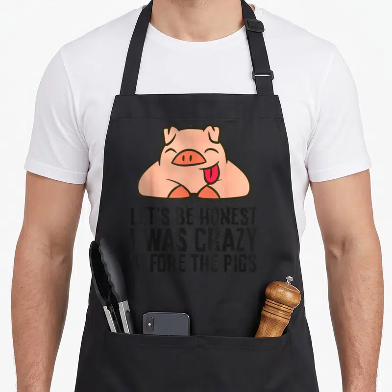 Honest crazy pig man apron - Pink cartoon pig above black text LET'S BE HONEST I WAS CRAZY BEFORE THE PIGS — shown on a black cotton canvas apron against a white background