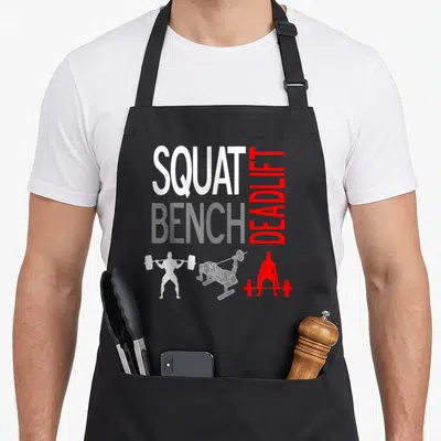 Squat bench apron - Stacked SQUAT BENCH DEADLIFT text above three weightlifter silhouettes in white, red, and grey — shown on a black cotton canvas apron against a white background