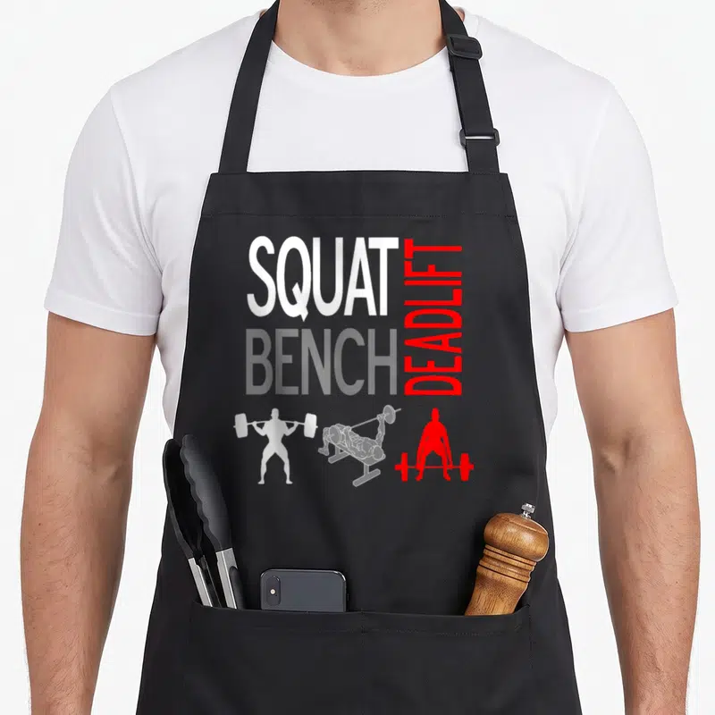 Squat bench apron - Stacked SQUAT BENCH DEADLIFT text above three weightlifter silhouettes in white, red, and grey — shown on a black cotton canvas apron against a white background