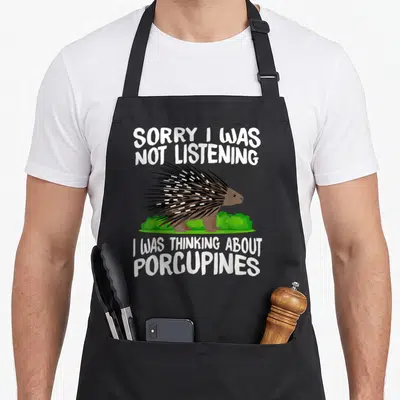Porcupine conservation animal apron - Cartoon brown porcupine with white quills on green bushes and white text — shown on a black cotton canvas apron against a white background