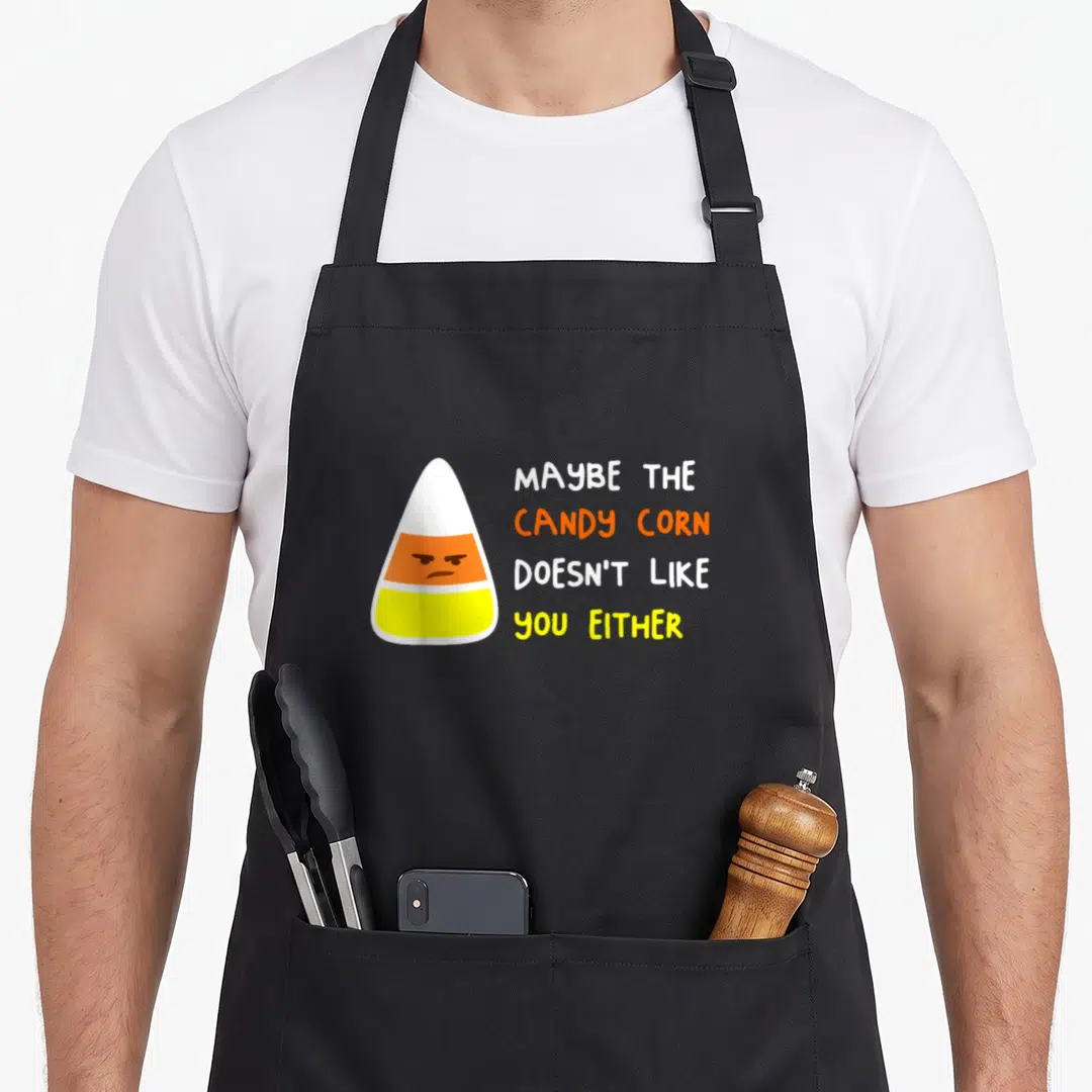 Costume candy halloween apron - Grumpy cartoon candy corn with white orange yellow text stacked right — shown on a black cotton canvas apron against a white background