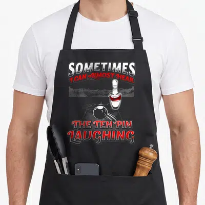 Laughing pins for aprons - Cartoon white bowling pin laughing with red mouth and grey ball under split text — shown on a black cotton canvas apron against a white background