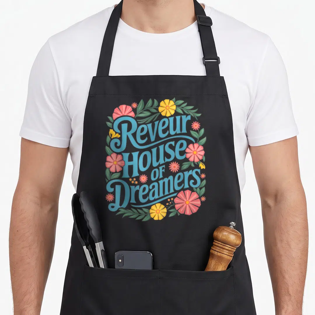 School dreamer apron - Stacked blue text Reveur House OF Dreamers framed by pink yellow and green floral wreath — shown on a black cotton canvas apron against a white background