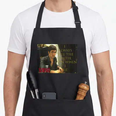 Montana scaredy apron - Photo of man sitting with gold stacked quote text and red Scarface logo on black background — shown on a black cotton canvas apron against a white background