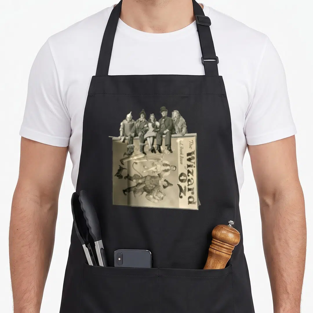 Scarecrow dorothy apron - Five Wizard of Oz characters sit atop book cover with vertical title text in sepia tones — shown on a black cotton canvas apron against a white background