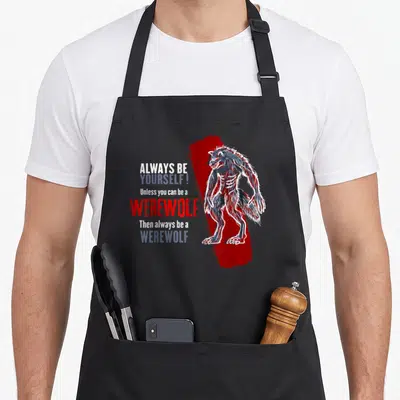Alway werewolf apron - Stacked text reading Always Be Werewolf next to a blue-grey cartoon wolf on a red paint stroke — shown on a black cotton canvas apron against a white background