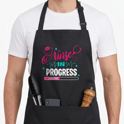 Progress nursing student apron - Nurse in Progress Loading text with pink stethoscope heart and blue EKG lines — shown on a black cotton canvas apron against a white background