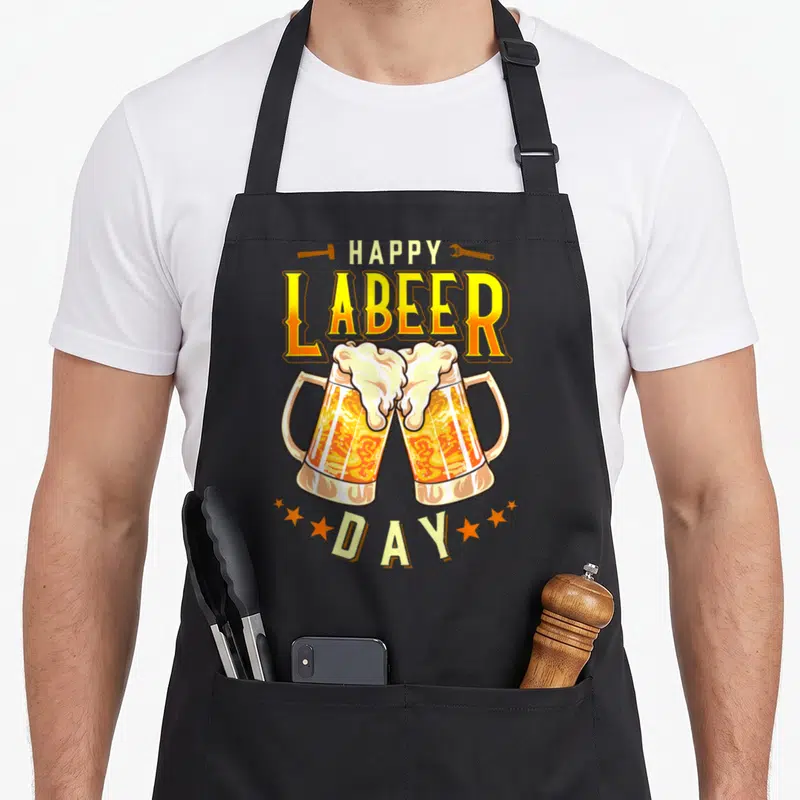 Labeer labor day apron - Two clinking beer mugs with HAPPY LABEER DAY text and tools in yellow, white, and orange — shown on a black cotton canvas apron against a white background