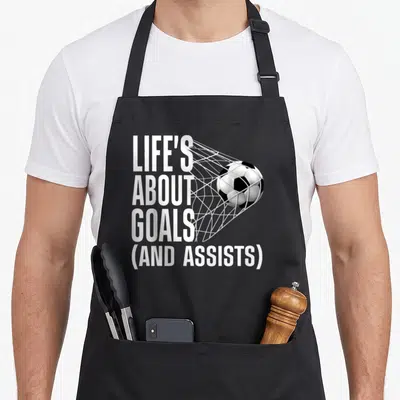 Goal football apron - Stacked white text LIFE'S ABOUT GOALS AND ASSISTS beside black and white soccer ball in goal net — shown on a black cotton canvas apron against a white background