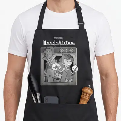 Funny cartoon apron - WandaVision text above retro TV with cartoon Wanda and Vision in grey white art style — shown on a black cotton canvas apron against a white background