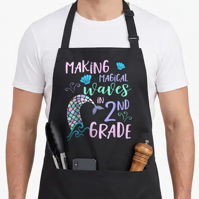 Grade 2nd apron - Making Magical Waves in 2nd Grade text with mermaid tail and seashells — shown on a black cotton canvas apron against a white background